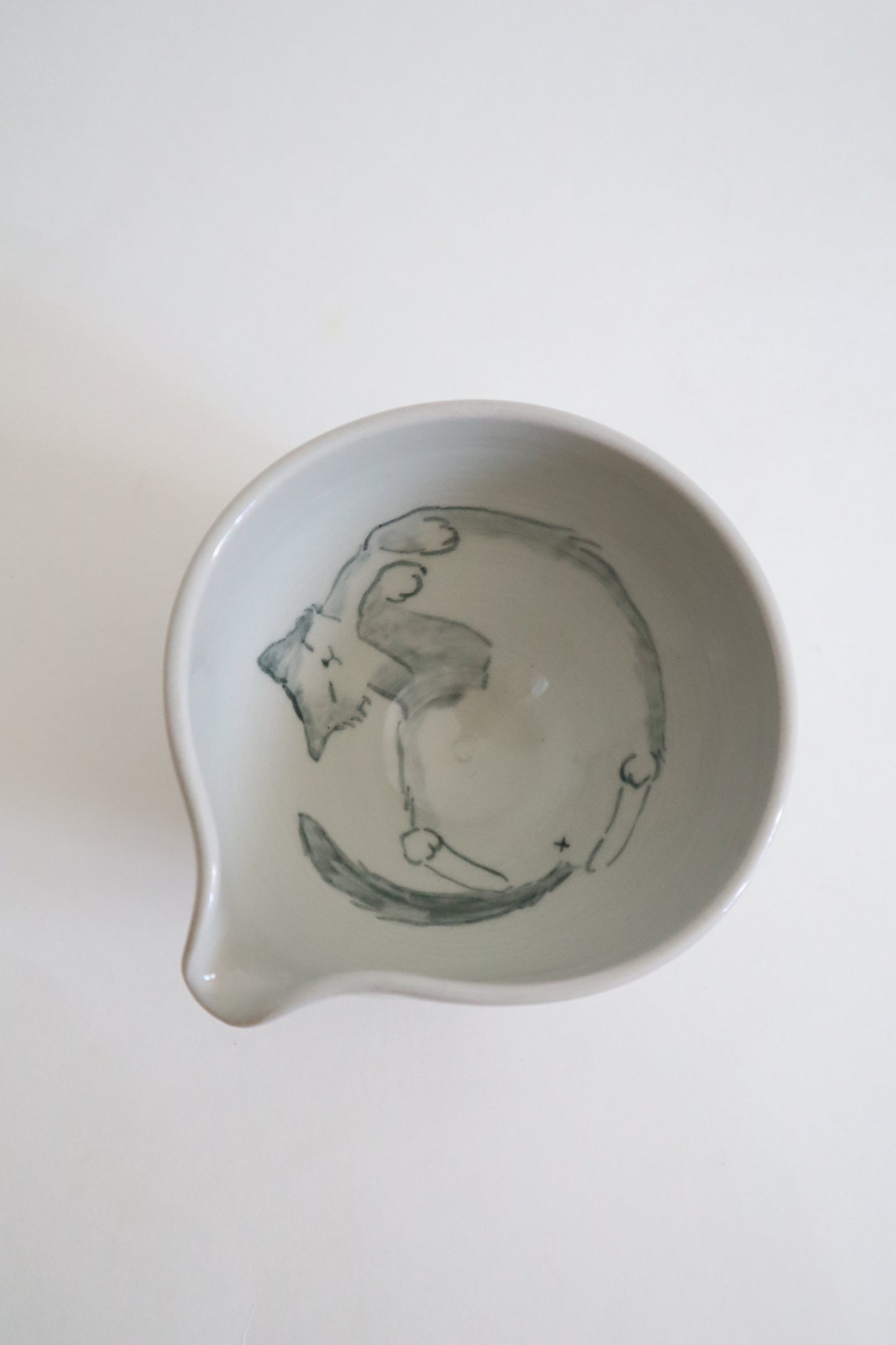 A white ceramic bowl with a small pouring spout, featuring a hand-painted gray illustration of a cat inside, shown from a top-down view on a white background.