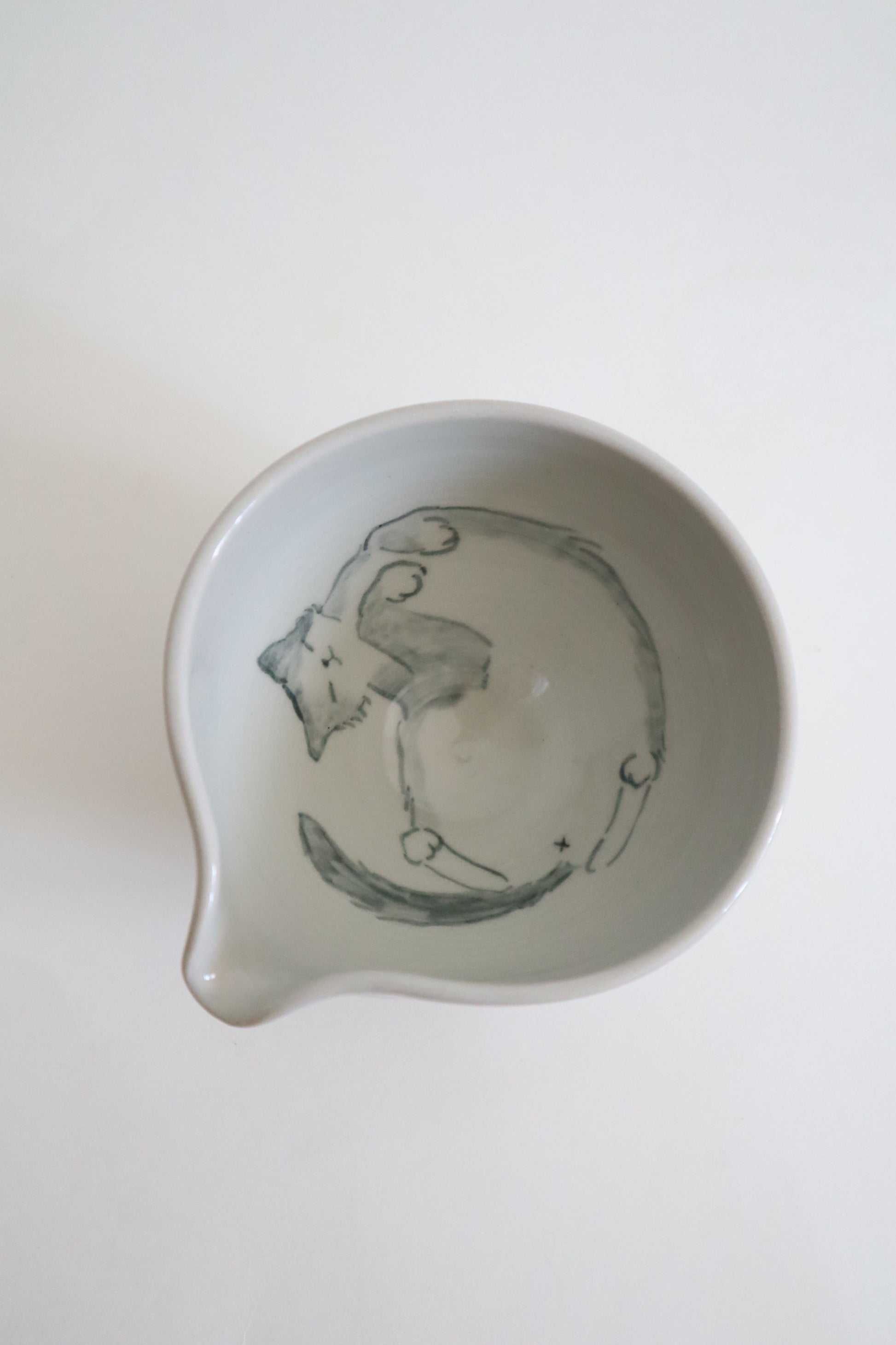 A white ceramic bowl with a small pouring spout, featuring a hand-painted gray illustration of a cat inside, shown from a top-down view on a white background.