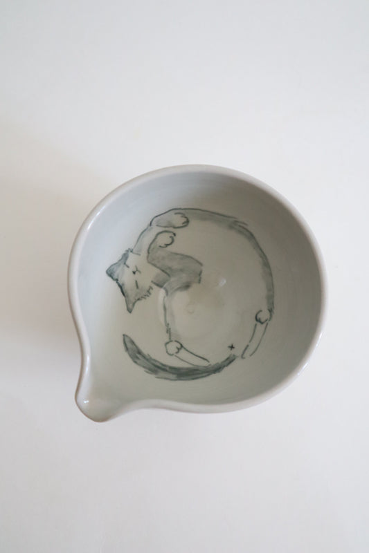 A white ceramic bowl with a small pouring spout, featuring a hand-painted gray illustration of a cat inside, shown from a top-down view on a white background.