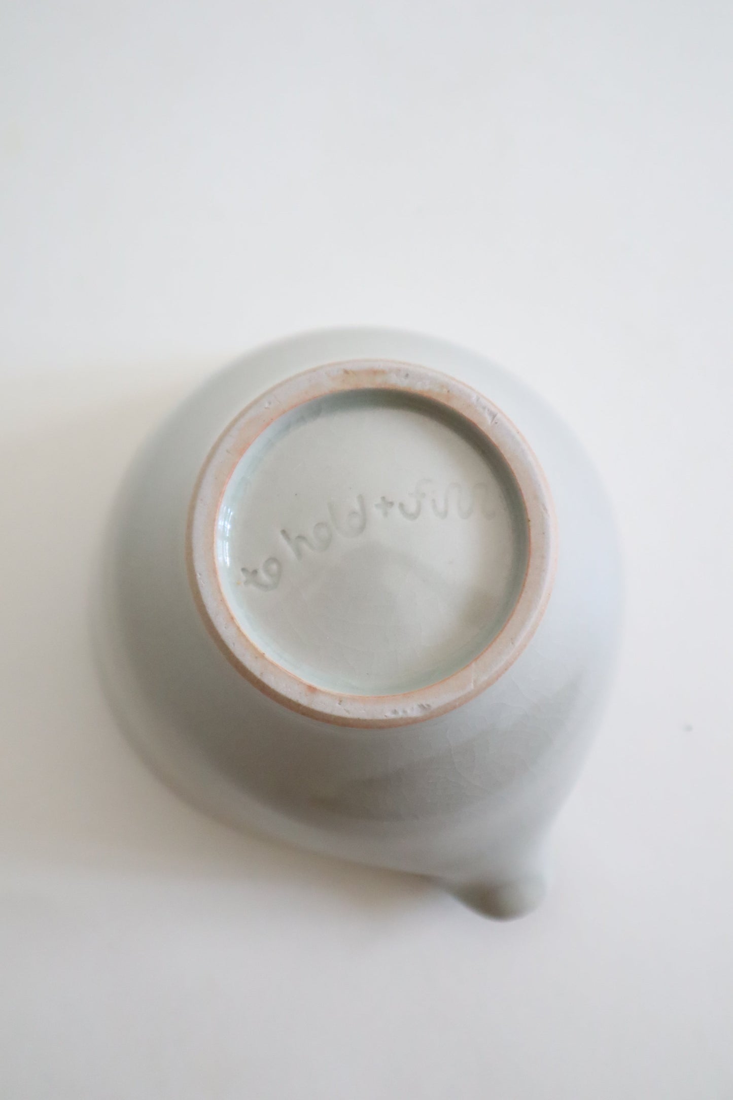 A top-down view of a matte ceramic teapot or pitcher with a small spout, showing its light gray glazed exterior and a natural beige clay rim around the base.