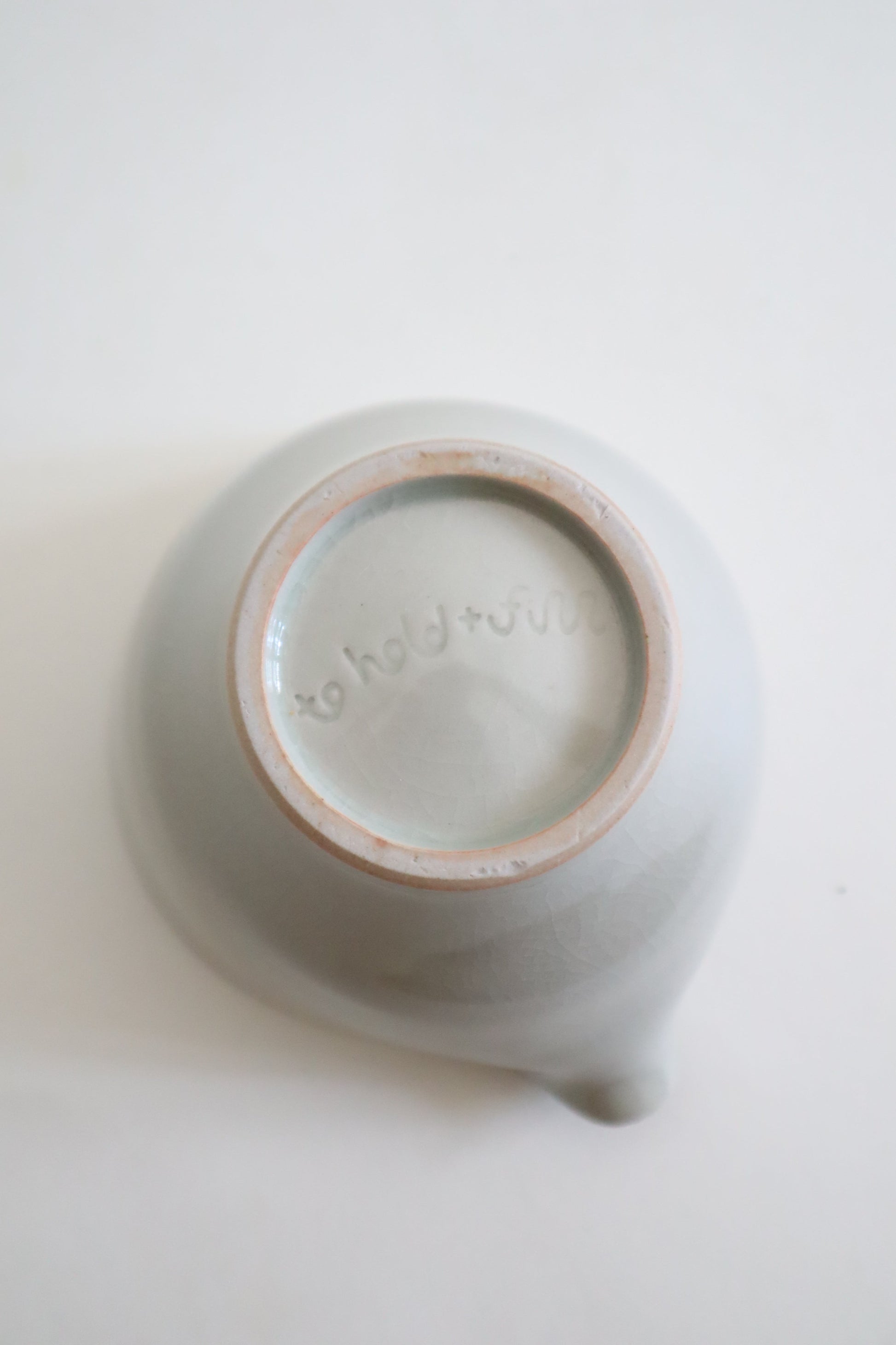 A top-down view of a matte ceramic teapot or pitcher with a small spout, showing its light gray glazed exterior and a natural beige clay rim around the base.