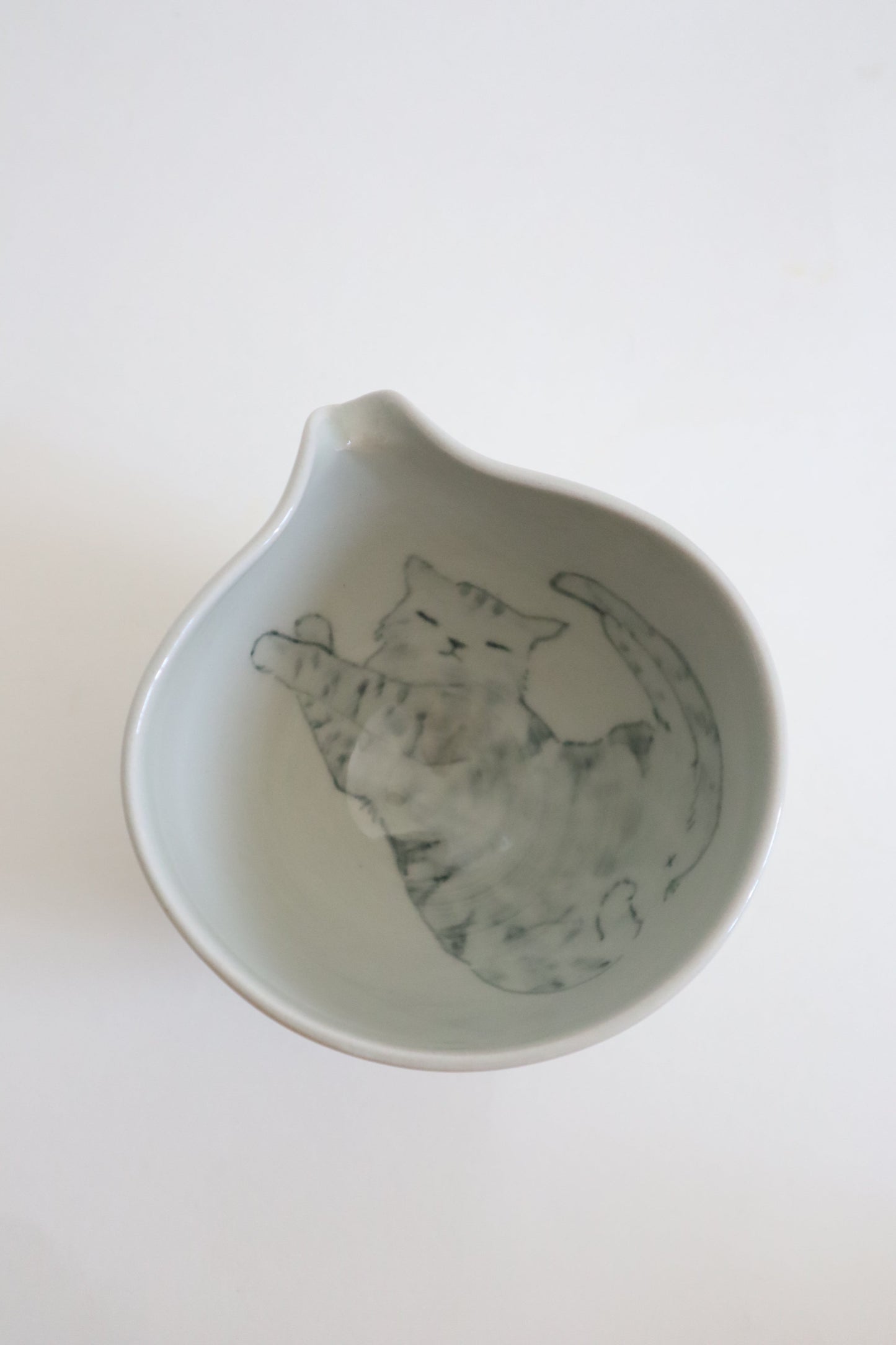 A light gray ceramic bowl with a small pouring spout, featuring a hand-painted illustration of a cat inside, shown against a plain white background.